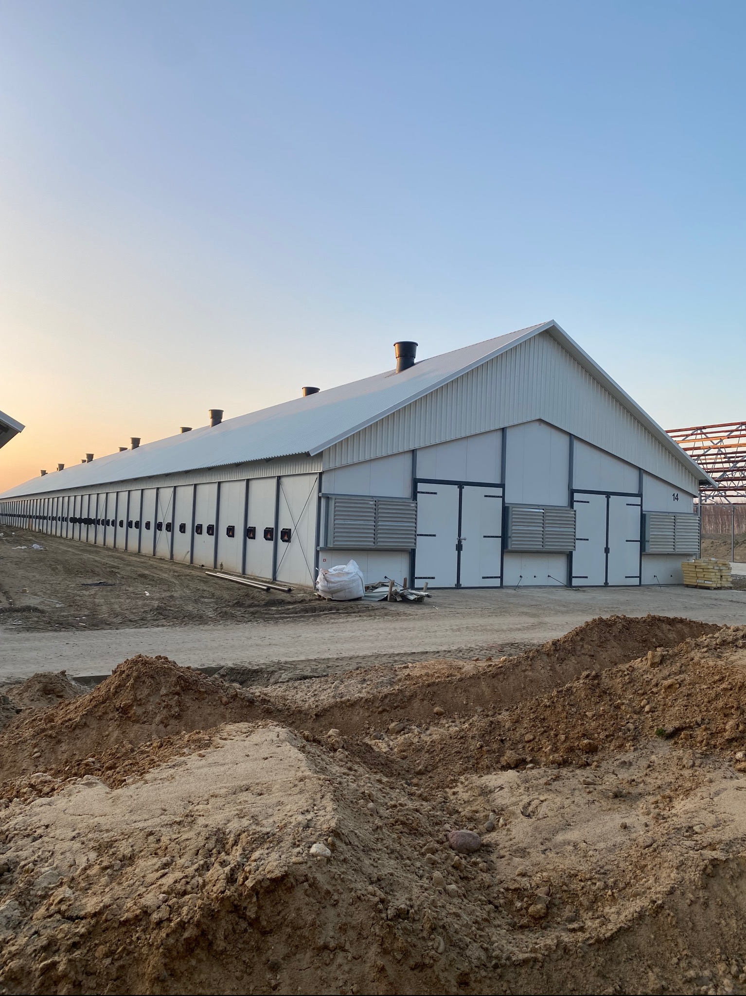 Długi, biały budynek gospodarczy z metalowym dachem i licznymi drzwiami, na tle błękitnego nieba o zmierzchu, z hałdami ziemi na pierwszym planie.