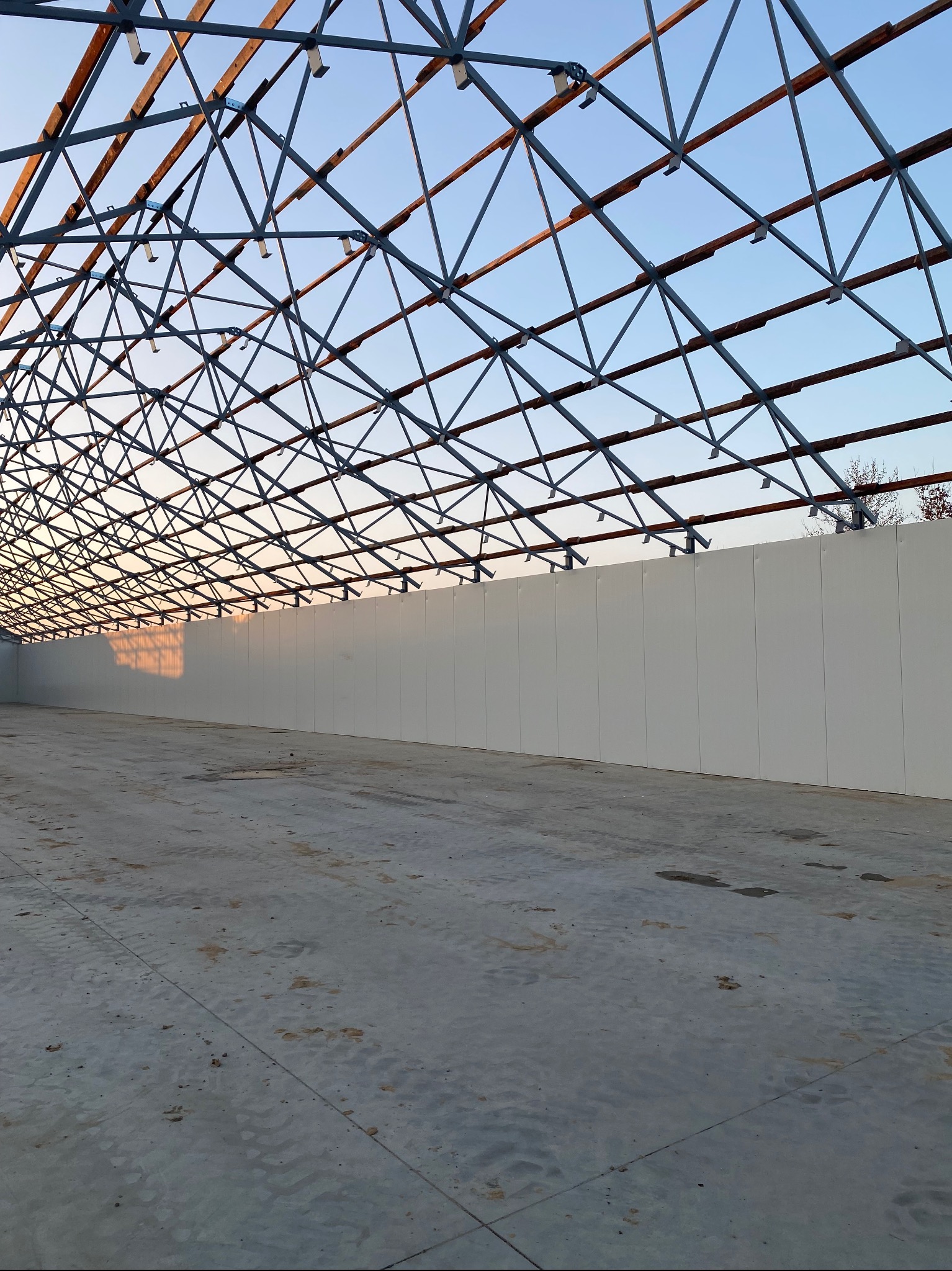 Konstrukcja stalowa dachu w hali produkcyjnej, widok od wewnątrz na szkielet dachu i betonową posadzkę. Widoczny fragment ściany bocznej hali.