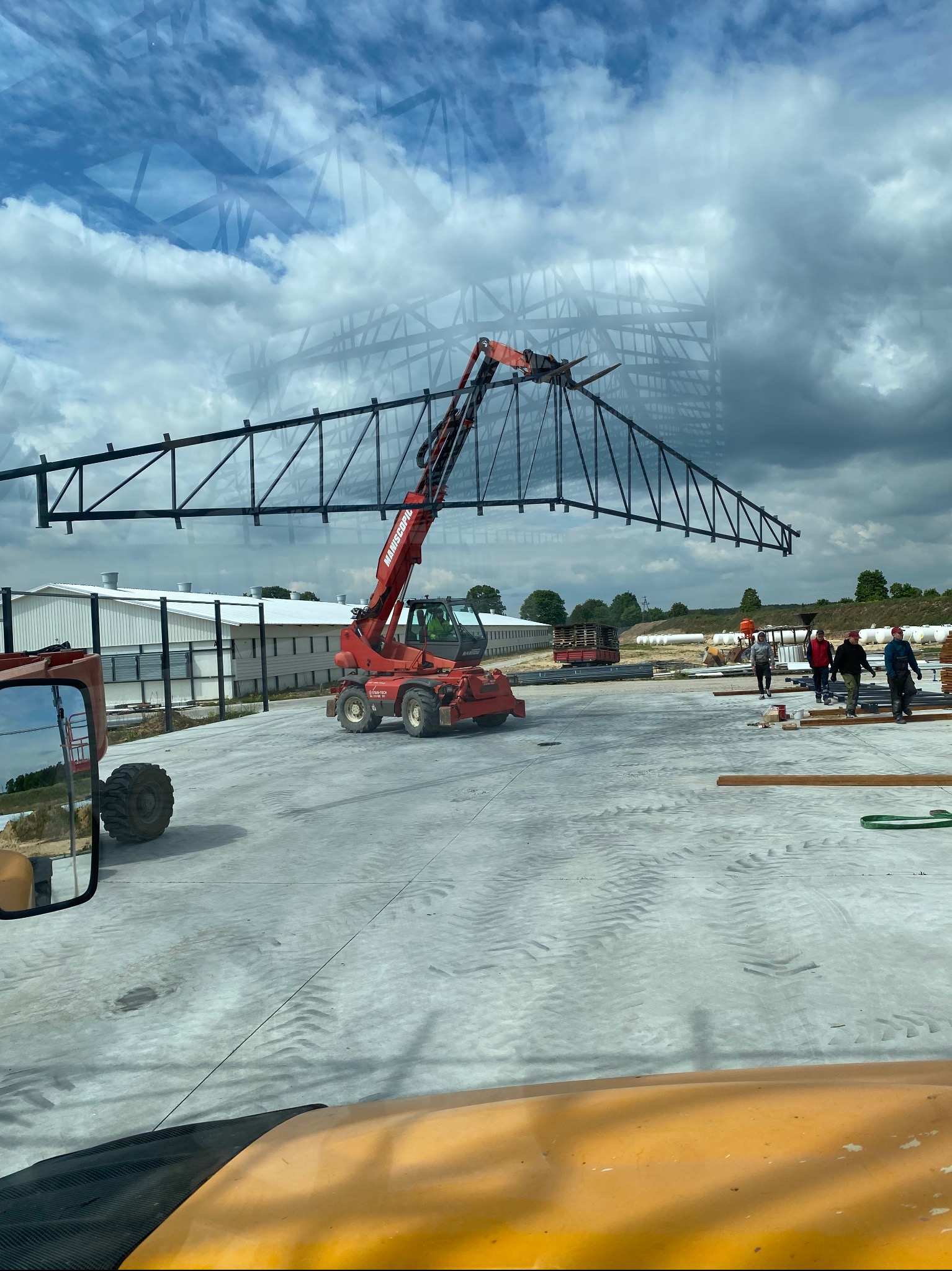 Montaż stalowej więźby dachowej dźwigiem Maniscopic na placu budowy. Widoczni pracownicy i elementy konstrukcyjne budynku. Ujęcie z kabiny pojazdu, odbicie nieba w szybie.