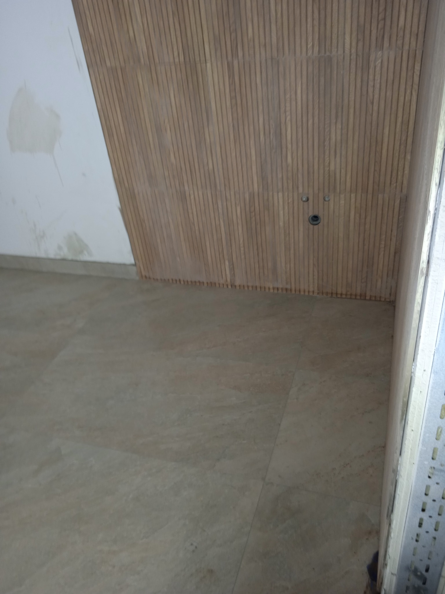 Wykończona ściana z drewnianych paneli pionowych w łazience, z widocznymi przyłączami. Na podłodze jasne płytki imitujące kamień. Fragment ściany przygotowany do malowania.