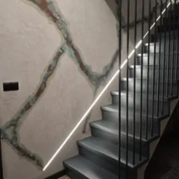 Nowoczesne schody z szarego drewna z minimalistyczną metalową balustradą i liniowym oświetleniem LED na ścianie z dekoracyjną fakturą imitującą rdzę.