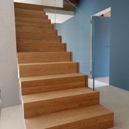 Zakład stolarski Mroczek - Drewniane schody z dębowymi stopniami i szklaną balustradą, błękitna ściana w tle, minimalistyczny design.