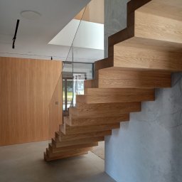 Zakład stolarski Mroczek - Nowoczesne, drewniane schody wspornikowe z balustradą ze szkła, przylegające do ściany o fakturze betonu architektonicznego. W tle widoczna drewniana boazeria i przeszklone drzwi.
