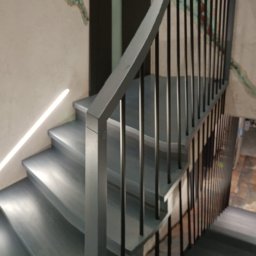 Zakład stolarski Mroczek - Nowoczesne schody z metalową balustradą i subtelnym oświetleniem LED wzdłuż ściany, wykończone w odcieniach szarości, z widoczną fakturą tynku na ścianie.