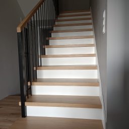 Zakład stolarski Mroczek - Schody drewniane z białymi podstopnicami i czarną metalową balustradą, widok z dołu, jasne oświetlenie.