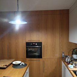 Zakład stolarski Mroczek - Kuchenna zabudowa z drewnianymi frontami, piekarnik wkomponowany w szafkę, blat z akcesoriami kuchennymi i niedojedzonym posiłkiem, oświetlenie punktowe.