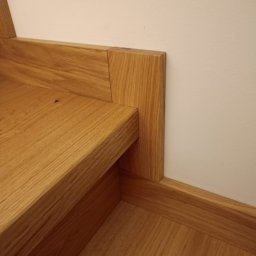 Zakład stolarski Mroczek - Detal wykończenia drewnianych schodów z widocznym połączeniem stopnia, podstopnicy i listwy przypodłogowej, wykonanych z jasnego drewna dębowego.