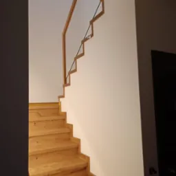 Drewniane schody z nietypową balustradą z drewnianych elementów połączonych czarnymi linkami, widok z dołu na białą ścianę i skos dachu z oknem.