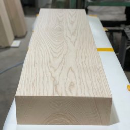 Zakład stolarski Mroczek - Surowy, jasny element z drewna o wyraźnym usłojeniu, leżący na stole roboczym z żółtą taśmą, w tle widoczna maszyna stolarska.