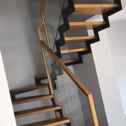 Zakład stolarski Mroczek - Nowoczesne schody drewniane z czarnymi metalowymi wspornikami i szklaną balustradą, widok z dołu, osoba w tle.