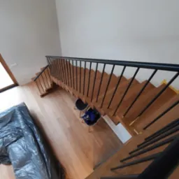 Widok z góry na drewniane schody z czarną balustradą, zabezpieczone folią malarską, z dwoma krzesłami w tle, na drewnianej podłodze.