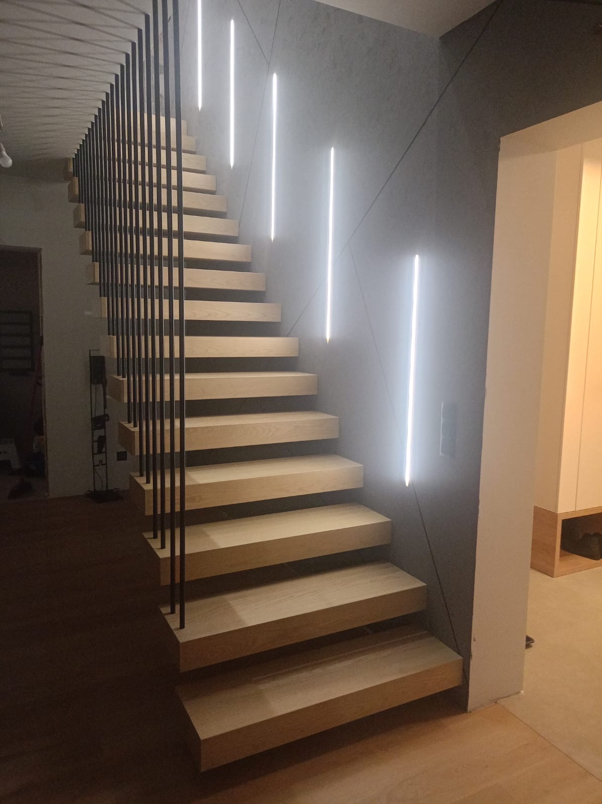 Nowoczesne schody drewniane z czarną metalową balustradą i pionowym oświetleniem LED na szarej ścianie.