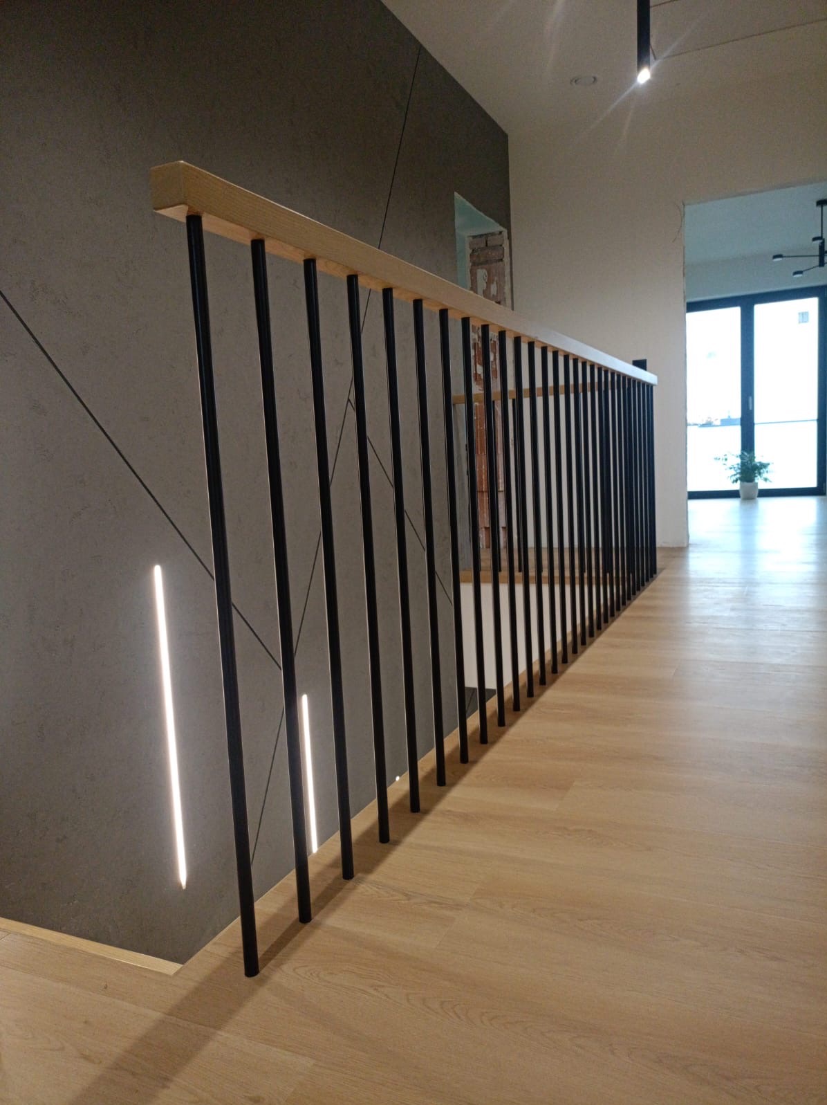 Nowoczesna balustrada schodowa z drewnianym poręczą i czarnymi metalowymi szczebelkami, oświetlenie LED w ścianie, panele podłogowe w odcieniach drewna.