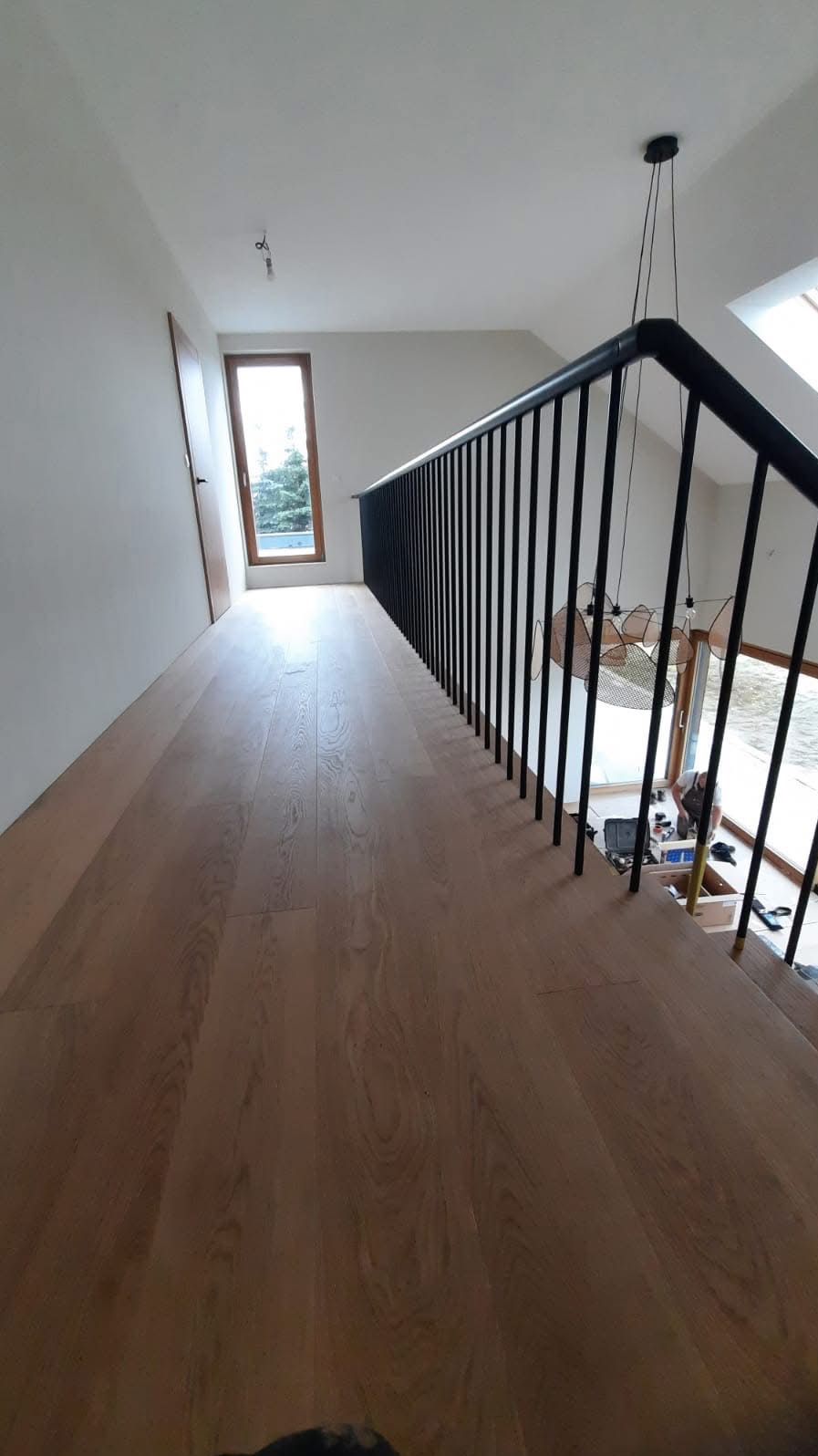 Jasny, drewniany parkiet na poddaszu z czarną balustradą, widoczne okno i drzwi w tle, oraz lampa wisząca.