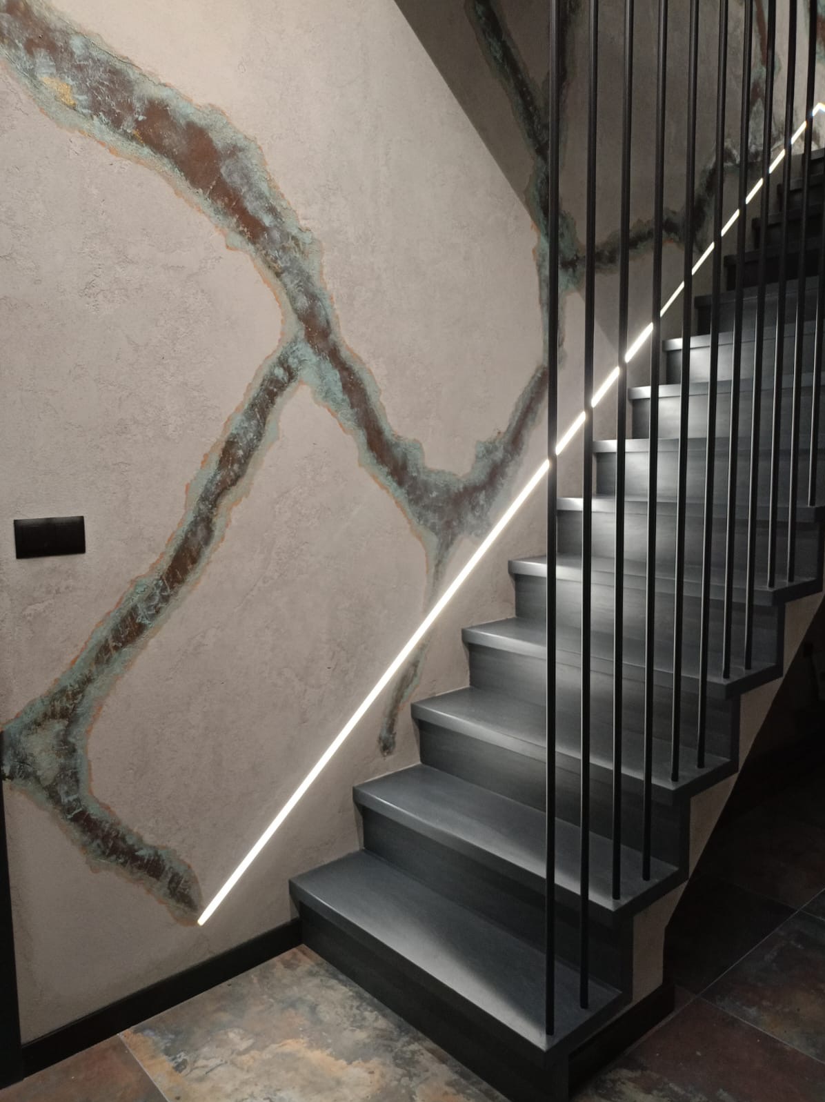 Nowoczesne schody z szarego drewna z minimalistyczną metalową balustradą i liniowym oświetleniem LED na ścianie z dekoracyjną fakturą imitującą rdzę.