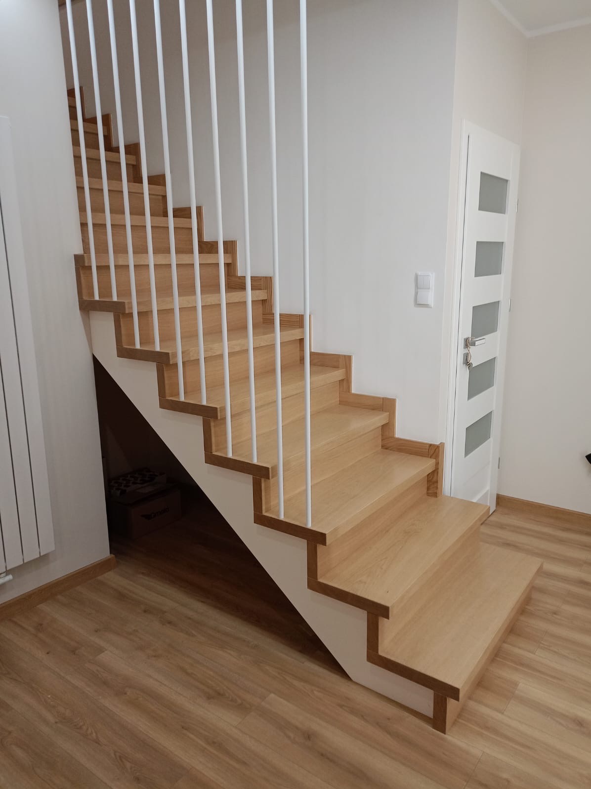 Drewniane schody z białymi, pionowymi balustradami w nowoczesnym wnętrzu, widok z dołu na pierwsze stopnie i część biegu schodów.