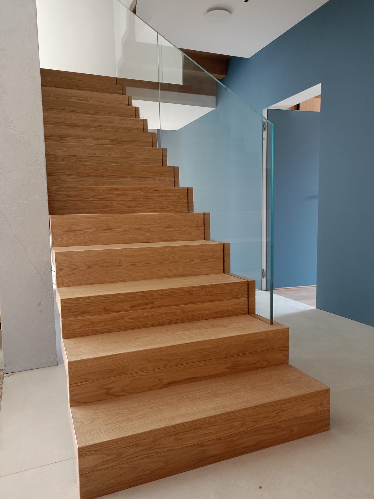 Drewniane schody z dębowymi stopniami i szklaną balustradą, błękitna ściana w tle, minimalistyczny design.