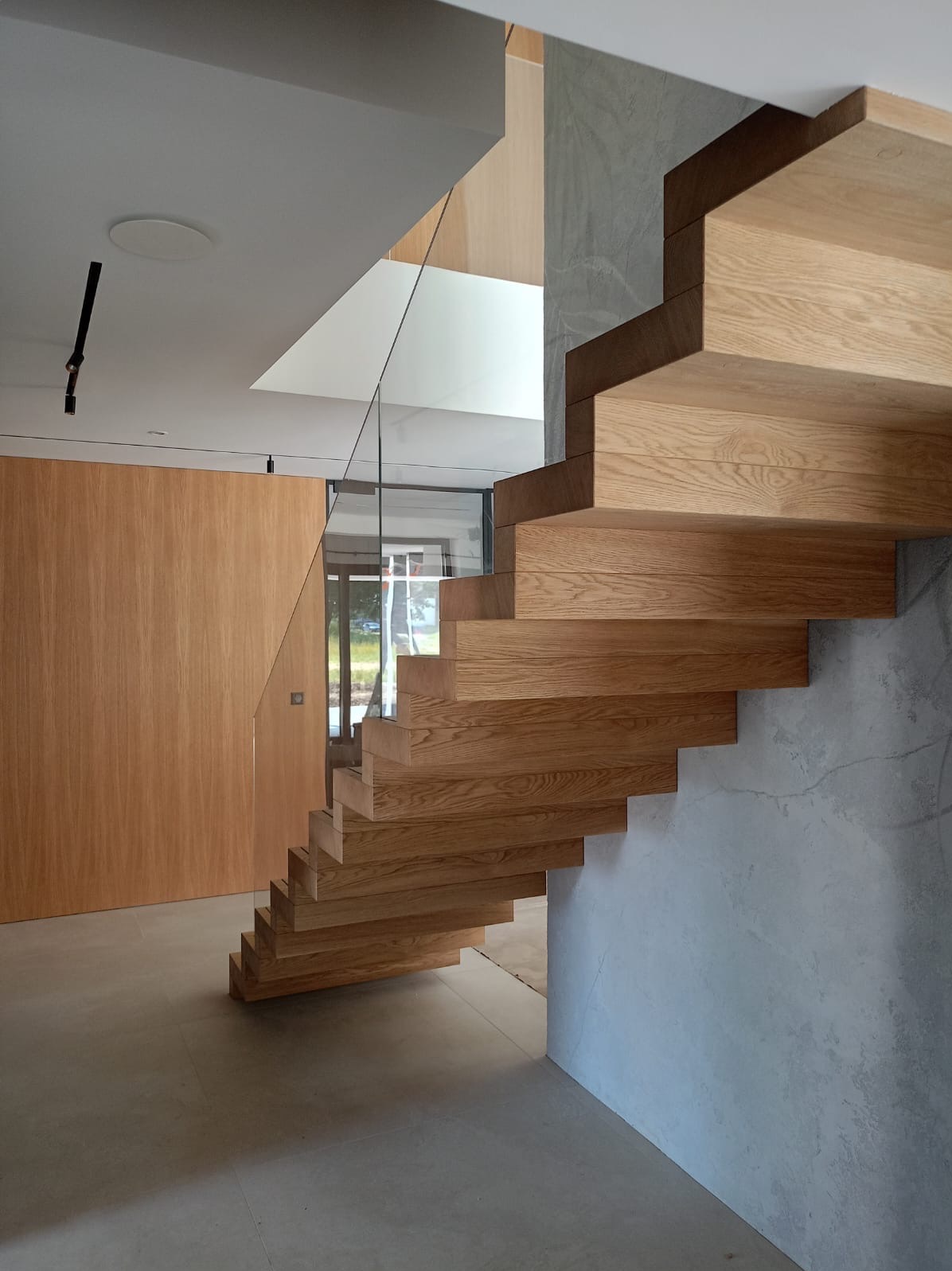 Nowoczesne, drewniane schody wspornikowe z balustradą ze szkła, przylegające do ściany o fakturze betonu architektonicznego. W tle widoczna drewniana boazeria i przeszklone drzwi.