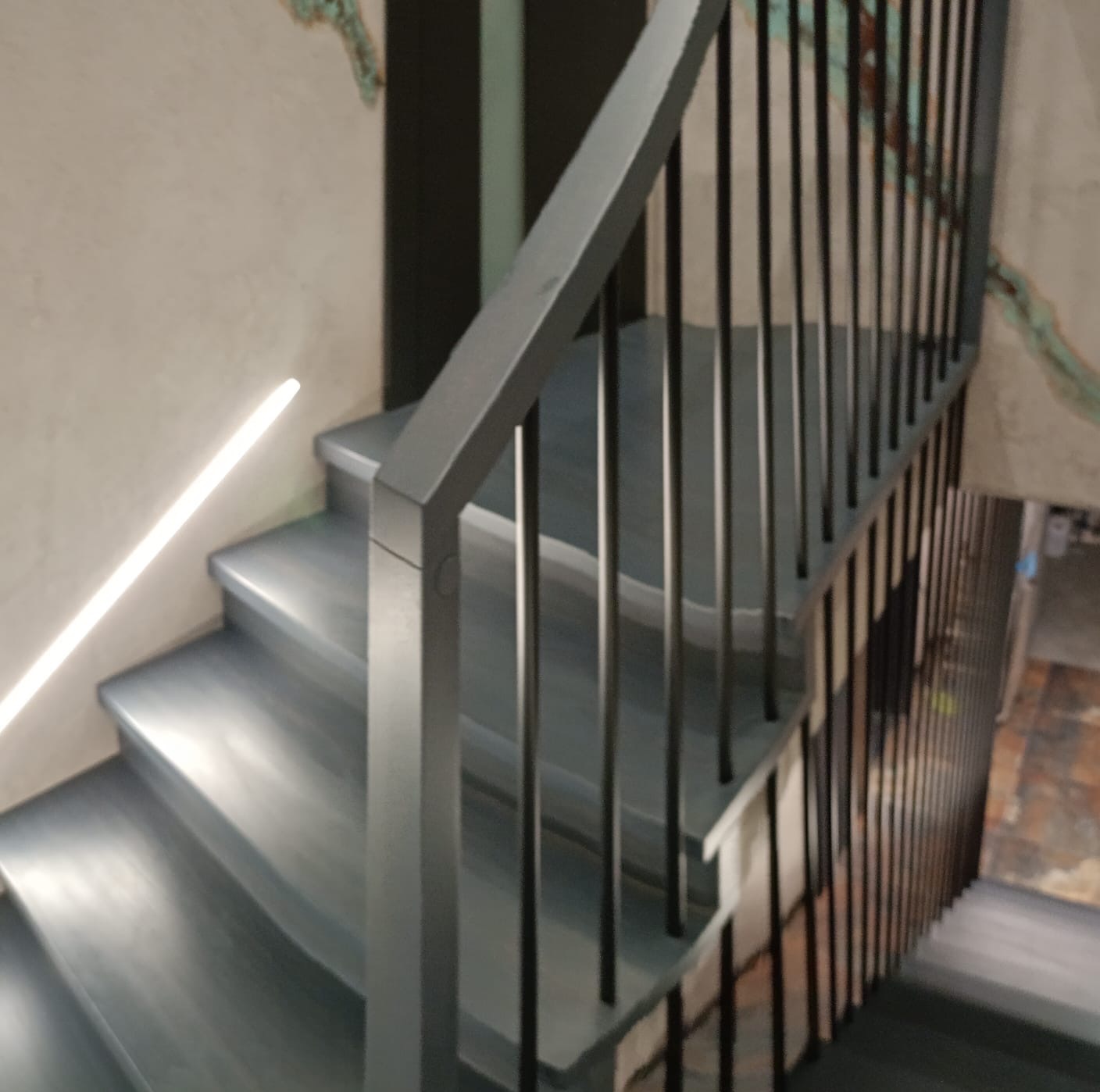 Nowoczesne schody z metalową balustradą i subtelnym oświetleniem LED wzdłuż ściany, wykończone w odcieniach szarości, z widoczną fakturą tynku na ścianie.