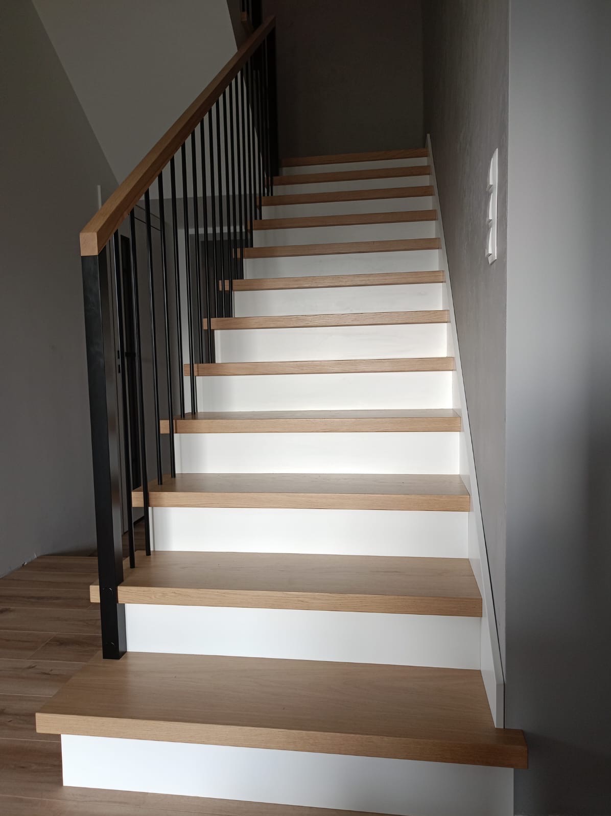 Schody drewniane z białymi podstopnicami i czarną metalową balustradą, widok z dołu, jasne oświetlenie.