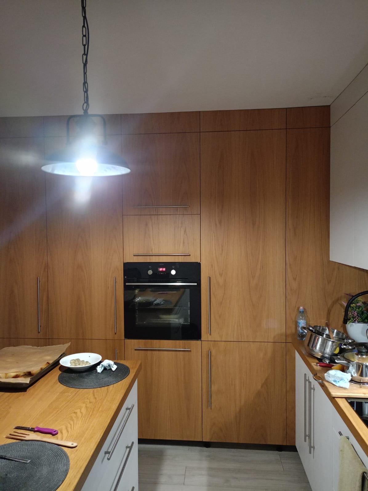 Kuchenna zabudowa z drewnianymi frontami, piekarnik wkomponowany w szafkę, blat z akcesoriami kuchennymi i niedojedzonym posiłkiem, oświetlenie punktowe.