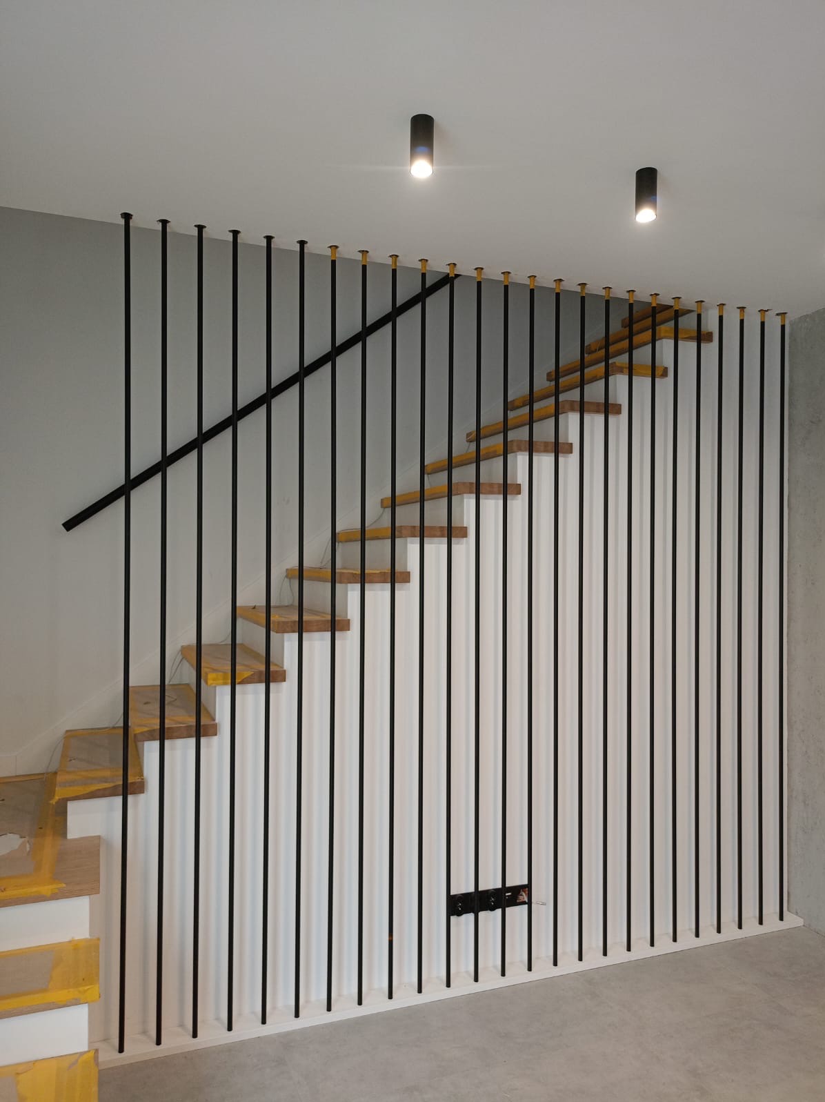 Drewniane schody z czarną balustradą z pionowych prętów, wykończone żółtą taśmą malarską na stopniach, oświetlone dwoma punktowymi lampami sufitowymi.