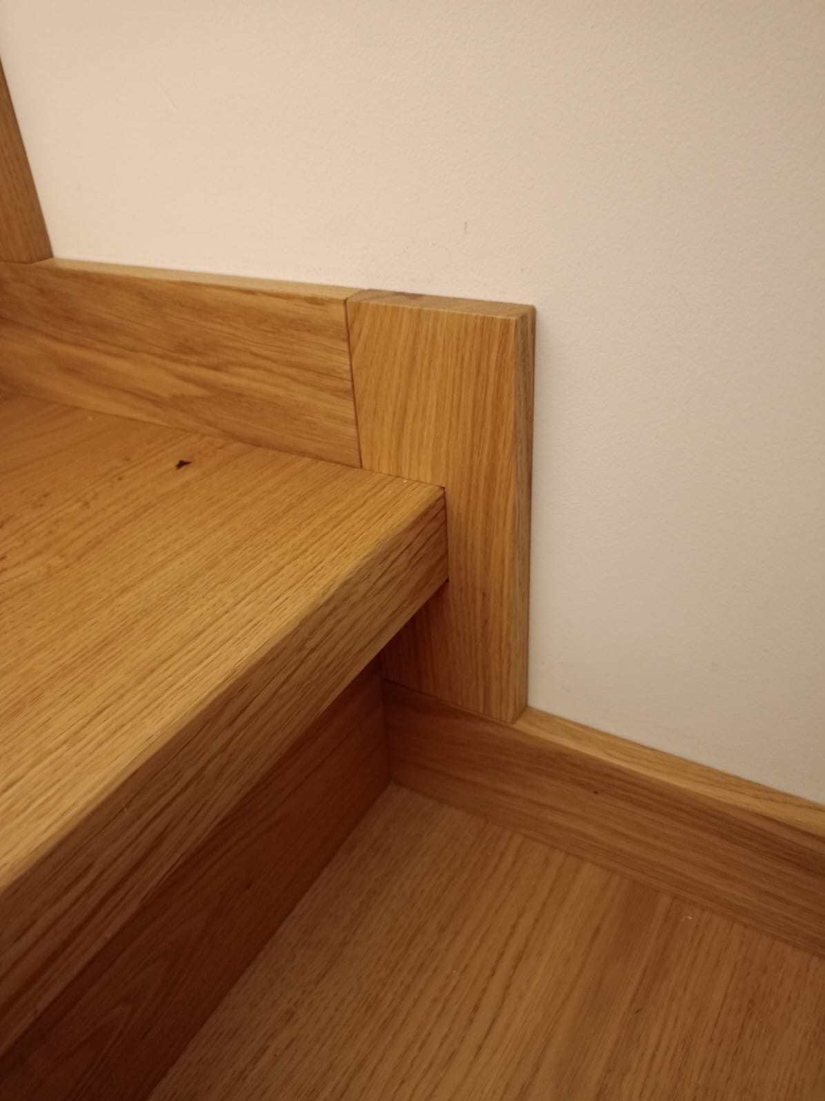 Detal wykończenia drewnianych schodów z widocznym połączeniem stopnia, podstopnicy i listwy przypodłogowej, wykonanych z jasnego drewna dębowego.