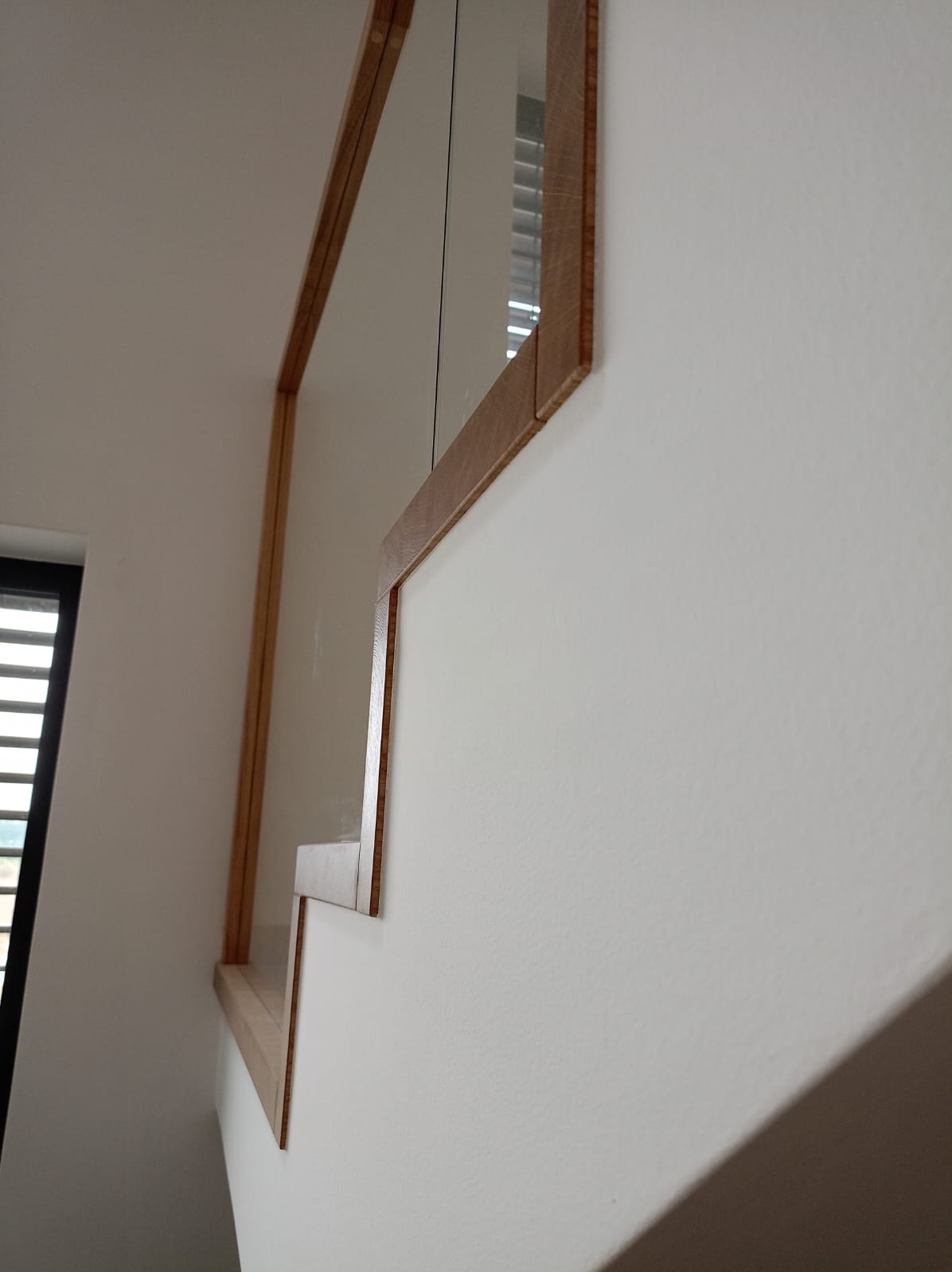 Nietypowe ujęcie fragmentu lustra w drewnianej ramie, przymocowanego do białej ściany. Rama wykonana z jasnego drewna, z widocznymi słojami. W lustrze odbija się okno z żaluzjami.