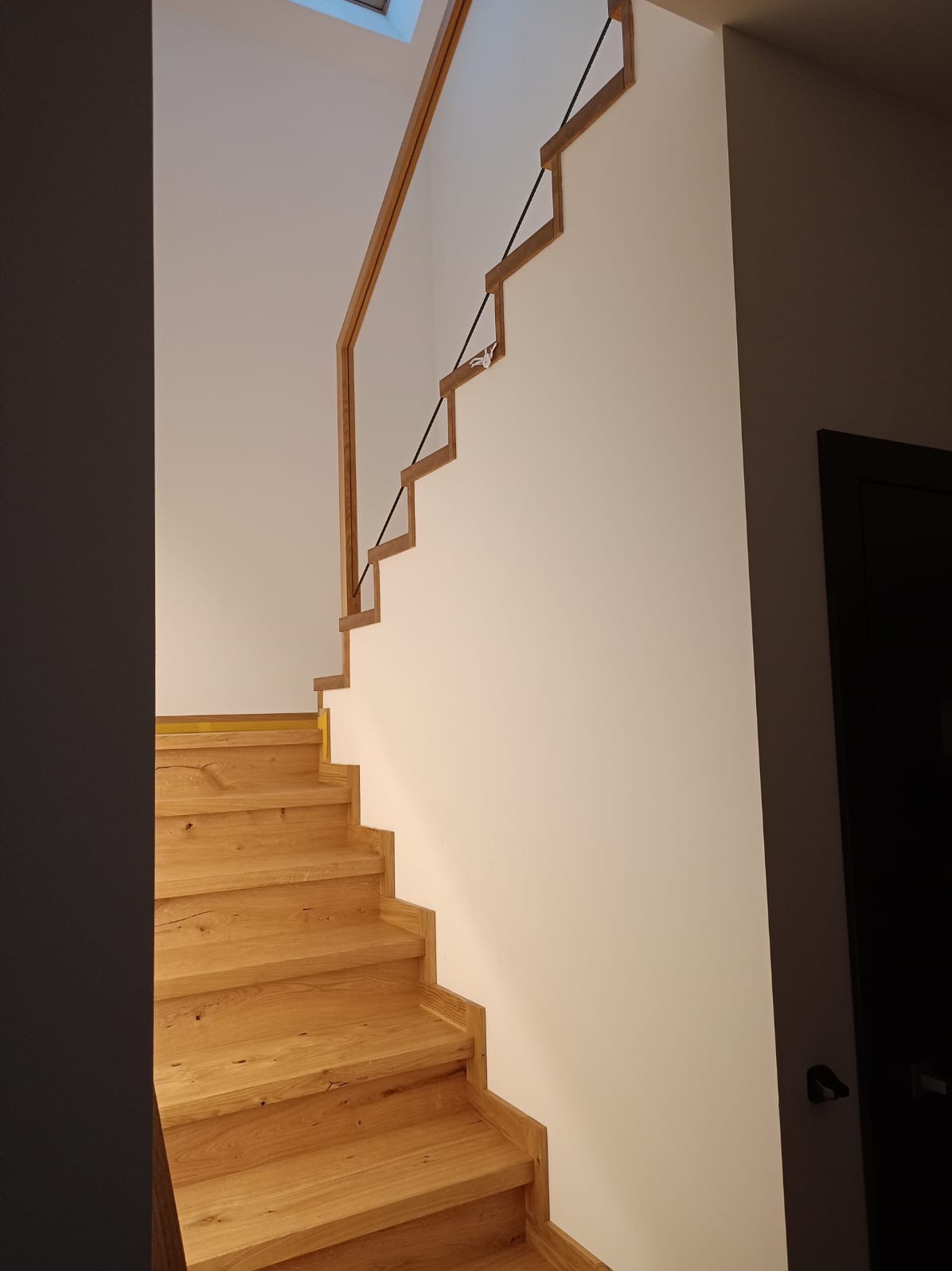Drewniane schody z nietypową balustradą z drewnianych elementów połączonych czarnymi linkami, widok z dołu na białą ścianę i skos dachu z oknem.