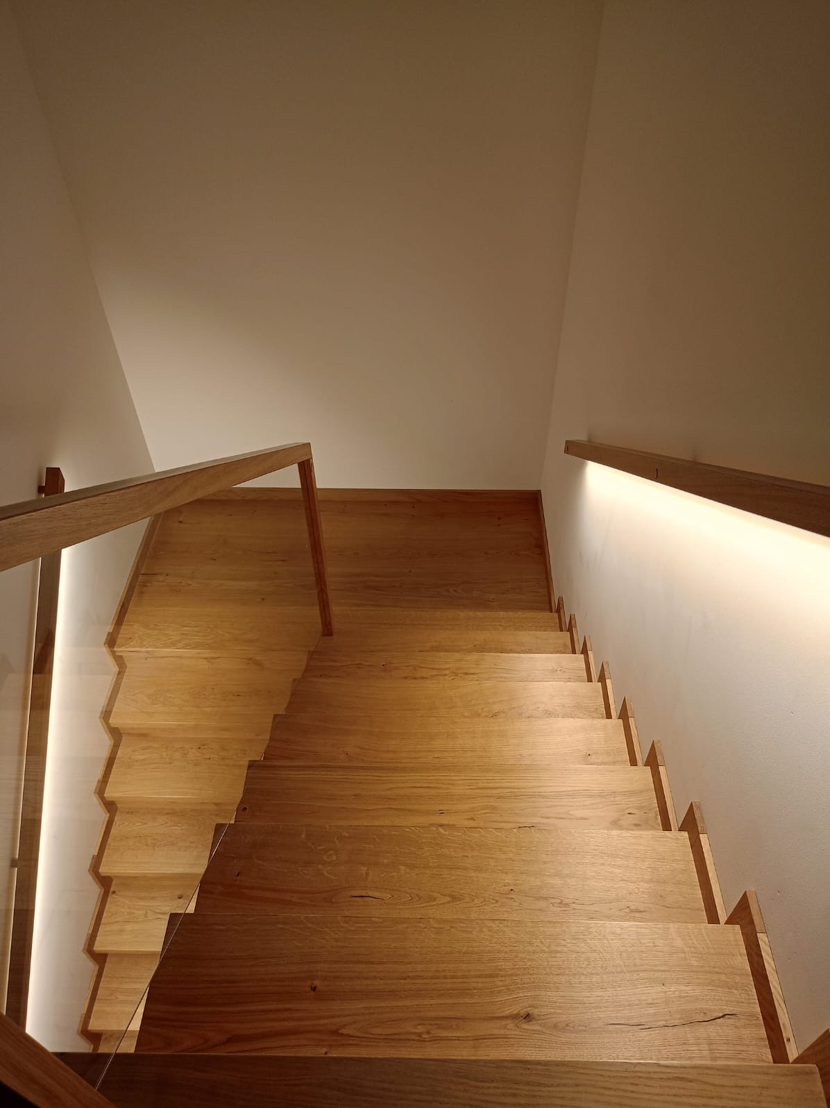 Drewniane schody z podświetleniem LED wzdłuż ściany i szklaną balustradą, widok z góry.