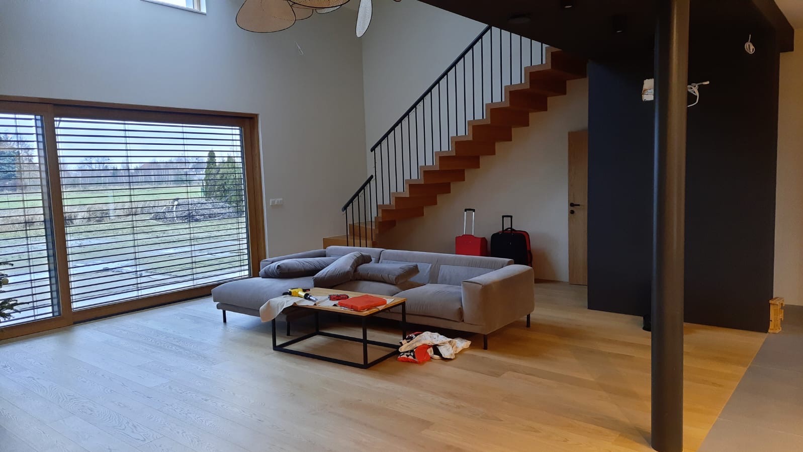 Jasna podłoga z desek w przestronnym salonie z szarą sofą i drewnianymi schodami na antresolę. W tle widoczne duże okno z żaluzjami i walizki podróżne.