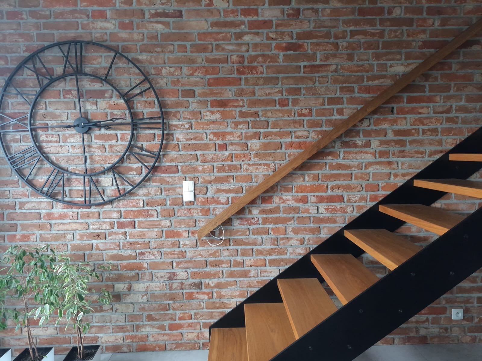 Wnętrze z ceglaną ścianą, dekoracyjnym zegarem, drewnianymi schodami z czarną konstrukcją i rośliną w doniczce.