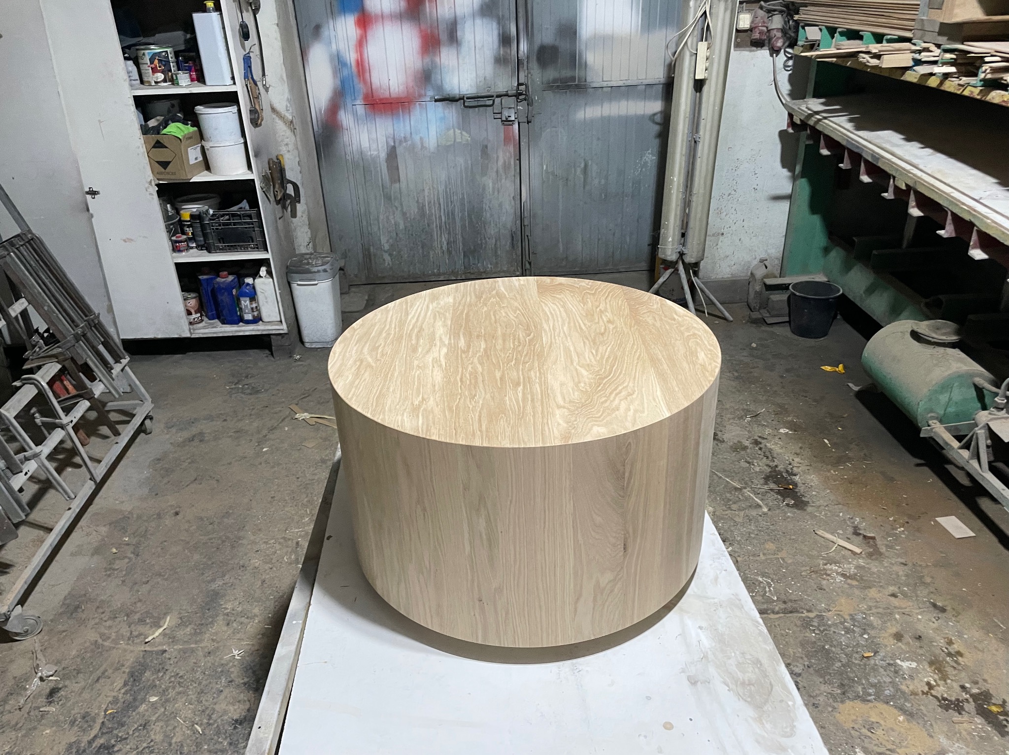 Okrągły blat drewniany o jasnym odcieniu w warsztacie stolarskim, widoczne drzwi warsztatu i regał z farbami