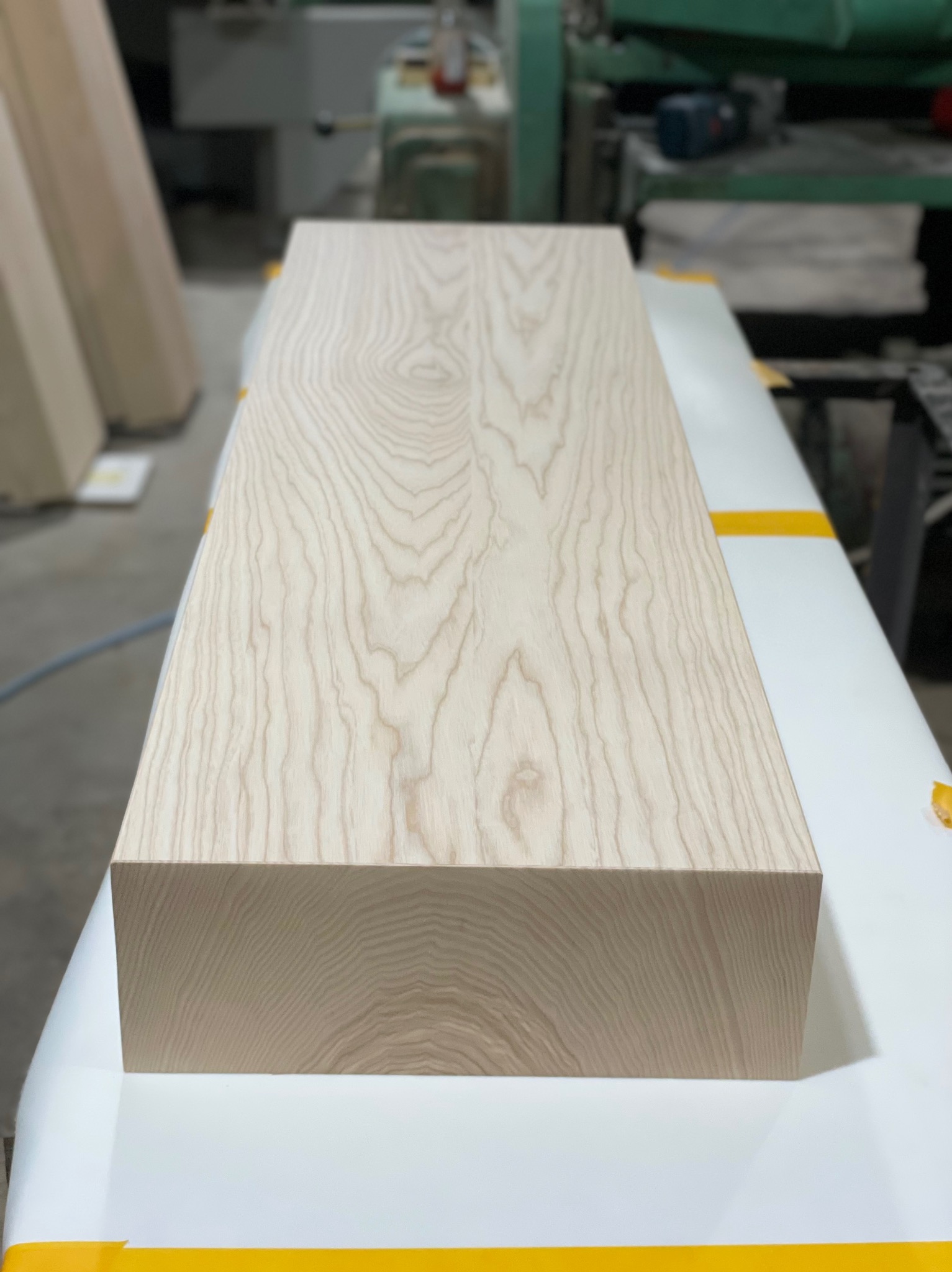 Surowy, jasny element z drewna o wyraźnym usłojeniu, leżący na stole roboczym z żółtą taśmą, w tle widoczna maszyna stolarska.