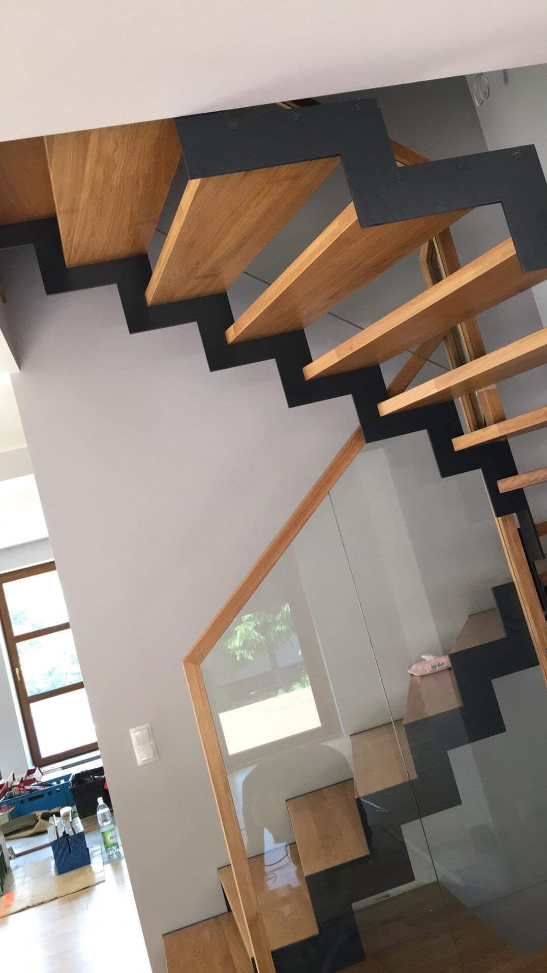 Nowoczesne schody drewniane z czarnymi wspornikami i szklaną balustradą, widok od dołu, z widocznym oknem w tle i elementami wyposażenia wnętrza.