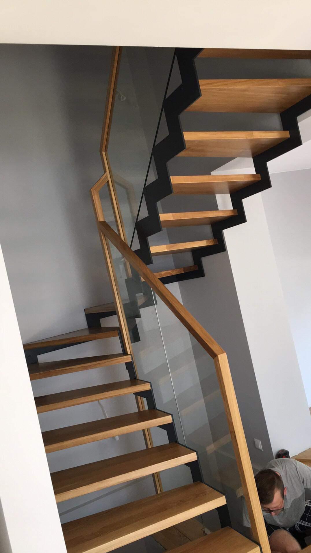Nowoczesne schody drewniane z czarnymi metalowymi wspornikami i szklaną balustradą, widok z dołu, osoba w tle.