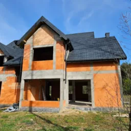 Dom w stanie surowym zamkniętym z czarną dachówką, widoczne rusztowanie i drabina, na tle błękitnego nieba i drzew.