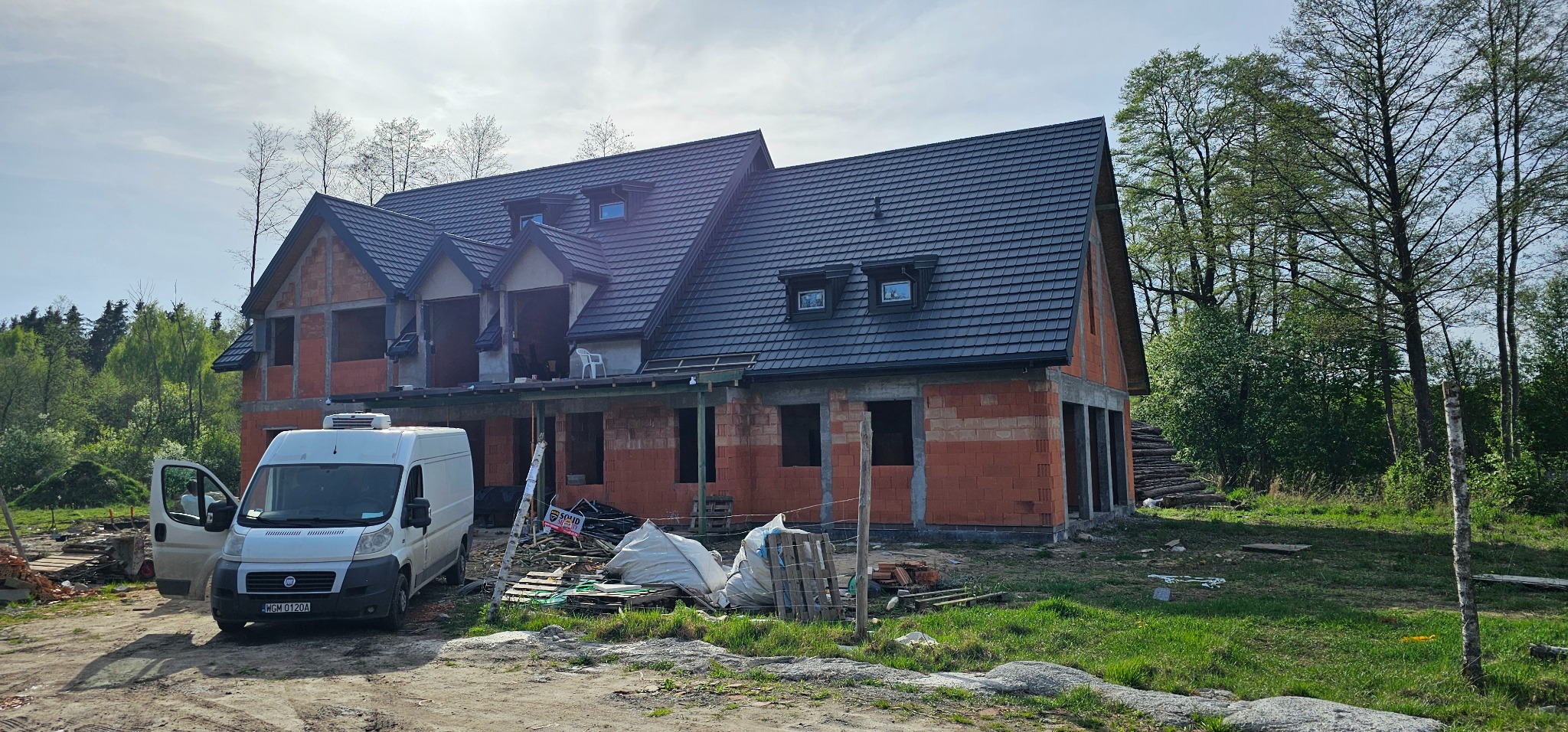 Dom w trakcie budowy z nowym, ciemnym dachem z lukarnami, biały samochód dostawczy na pierwszym planie, teren budowy z materiałami budowlanymi.