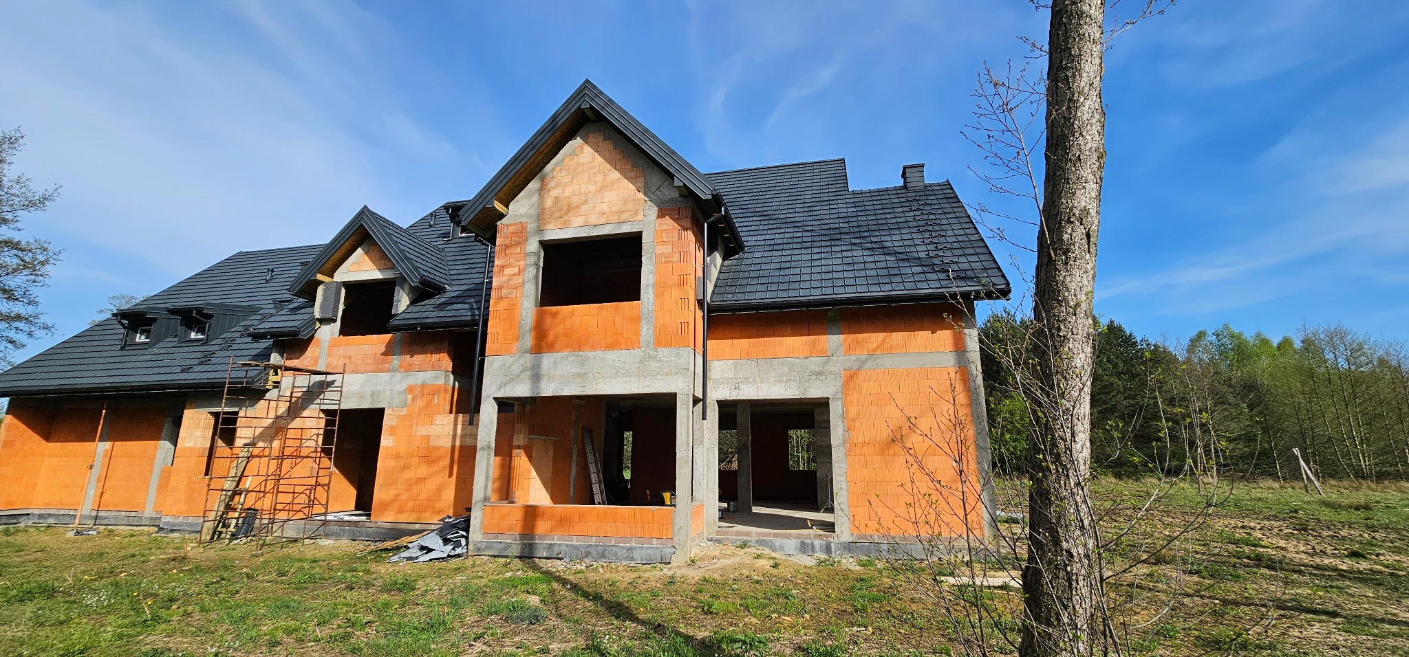 Dom w stanie surowym zamkniętym z czarną dachówką, widoczne rusztowanie i drabina, na tle błękitnego nieba i drzew.