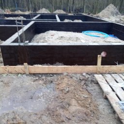 OM-GOR OLEH HORBYK - Wykop pod fundamenty z zalanymi ścianami, zabezpieczonymi izolacją przeciwwilgociową, widoczne zbrojenia i rury instalacyjne, na pierwszym planie drewniane szalunki i błotniste podłoże.