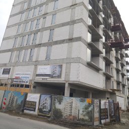 OM-GOR OLEH HORBYK - Wielopiętrowy budynek w stanie surowym, z widocznymi pustakami i szalunkami balkonowymi, zabezpieczony siatką z reklamami dewelopera.