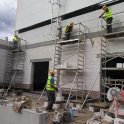 Czterech murarzy w żółtych kaskach i odblaskowych kamizelkach na rusztowaniach podczas wznoszenia ściany z białych bloczków betonu komórkowego Ytong; widoczna drabina przymocowana do ściany...