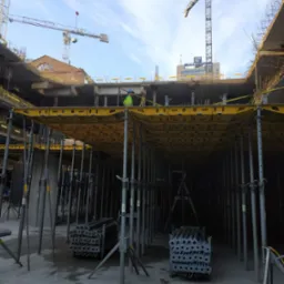 Wnętrze budowy z widoczną konstrukcją szalunkową pod przyszły strop, podpory stalowe, pracownik w odblaskowej kamizelce i żurawie budowlane w tle.