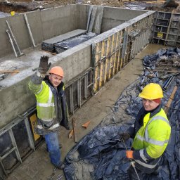 OM-GOR OLEH HORBYK - Dwóch robotników w kaskach i odblaskowych kurtkach przy budowie betonowego fundamentu, jeden macha ręką, widoczne szalunki i zbrojenia.