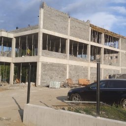 OM-GOR OLEH HORBYK - Wznoszenie dwukondygnacyjnego budynku z szarych bloczków, widoczne podpory stropu, zbrojenie, na górze budynku dwóch pracowników, obok zaparkowany czarny samochód osobowy.