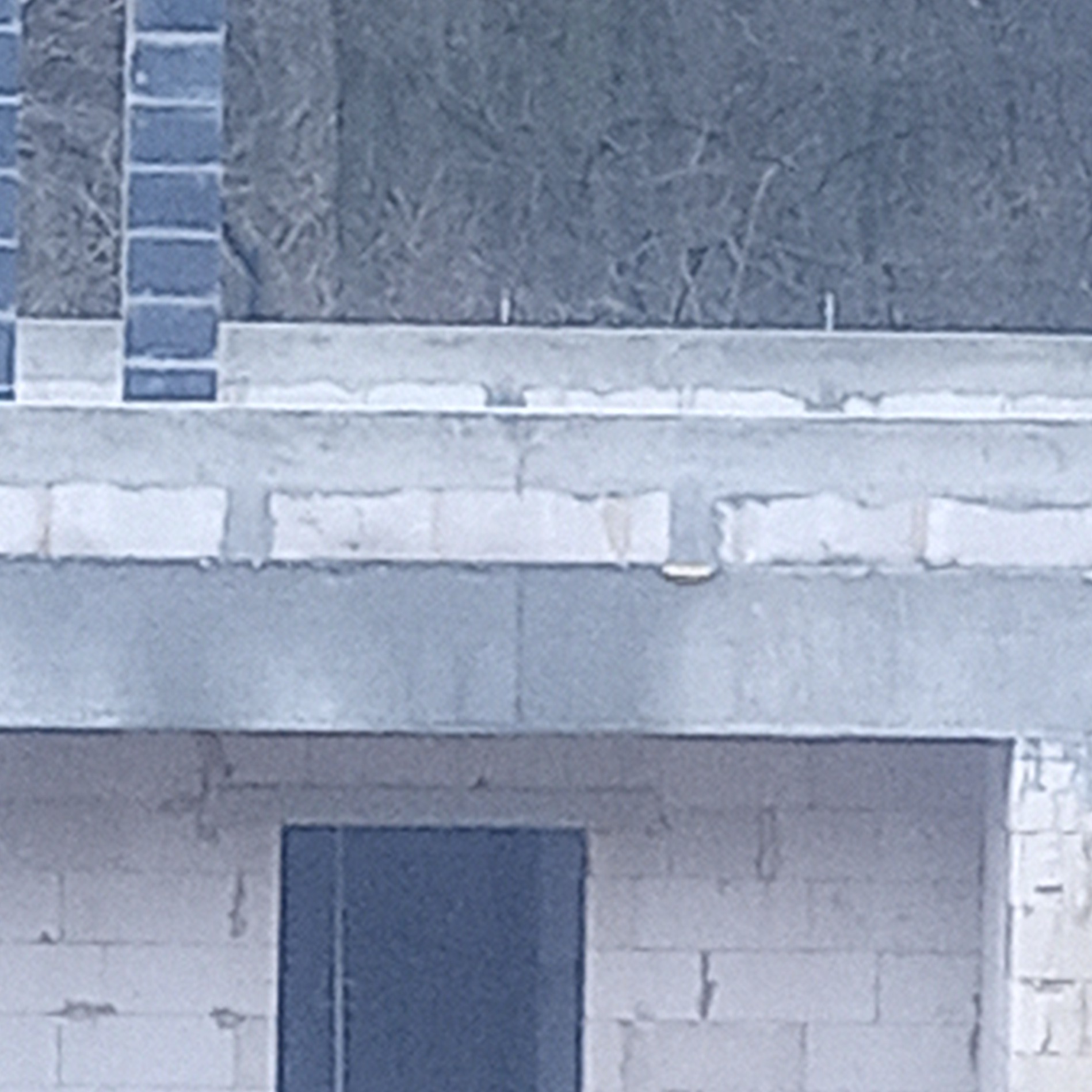 Fragment ściany budynku w trakcie murowania z betonu komórkowego, widoczne nadproże nad otworem okiennym/drzwiowym, w tle zarośla.