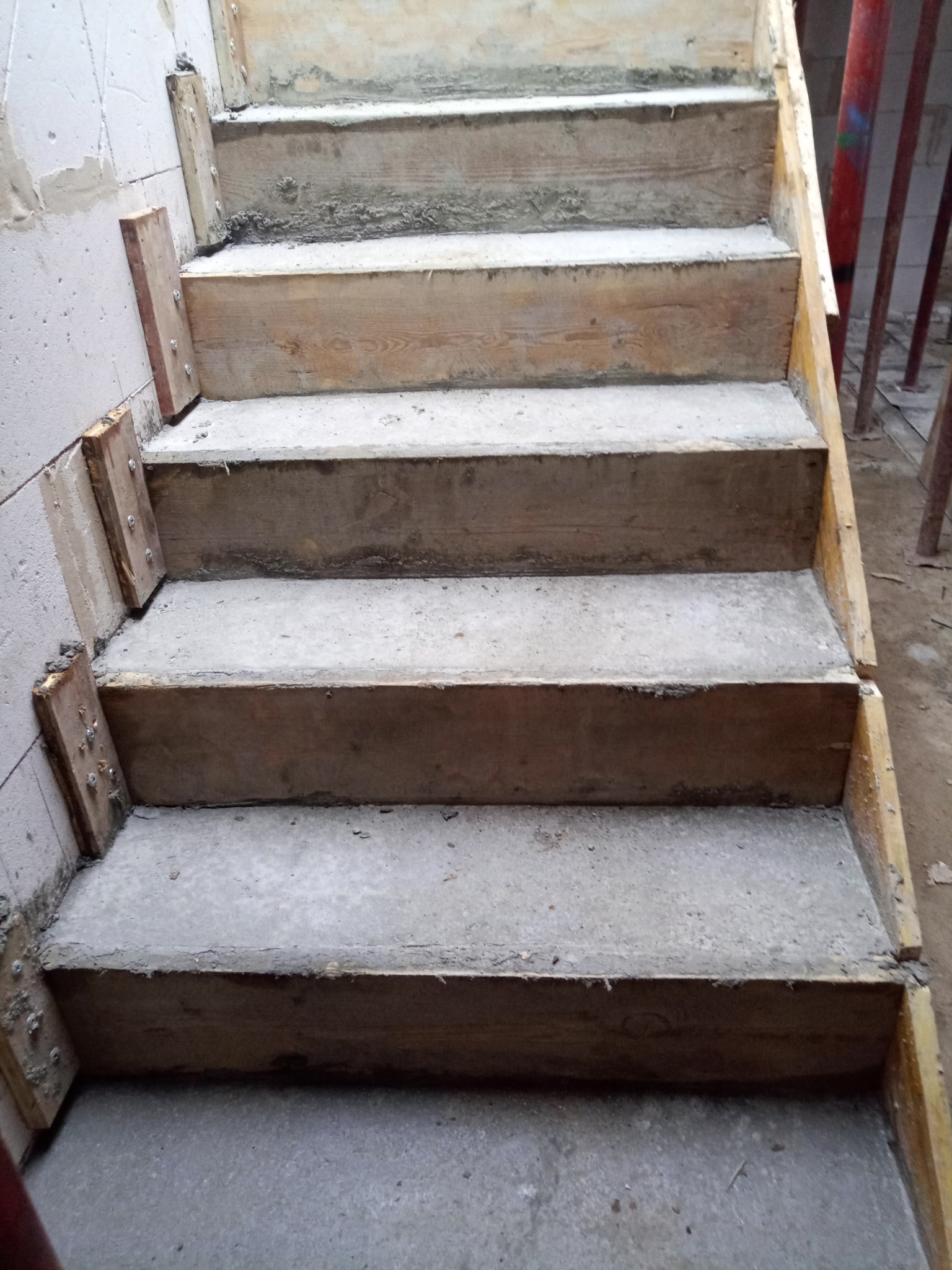Surowe betonowe schody w trakcie budowy, z drewnianym szalunkiem i widocznymi śladami betonu na ścianie.