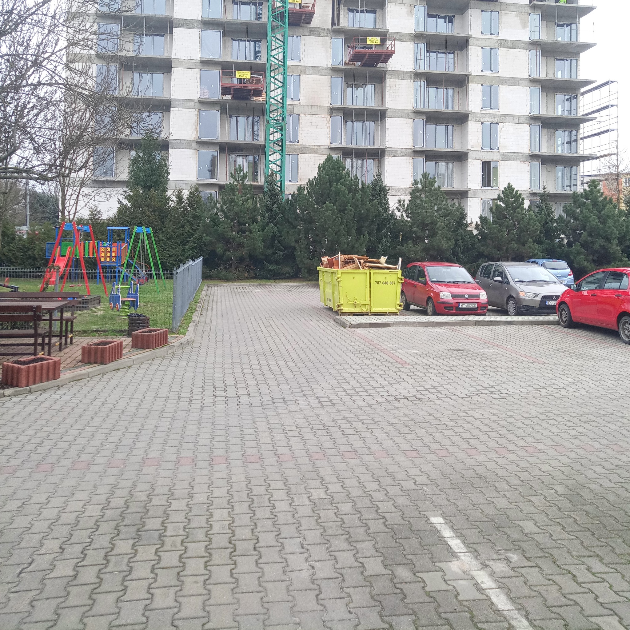 Wielopiętrowy budynek w stanie surowym z widocznym dźwigiem, kontener na odpady budowlane, parking z zaparkowanymi samochodami oraz plac zabaw w tle.