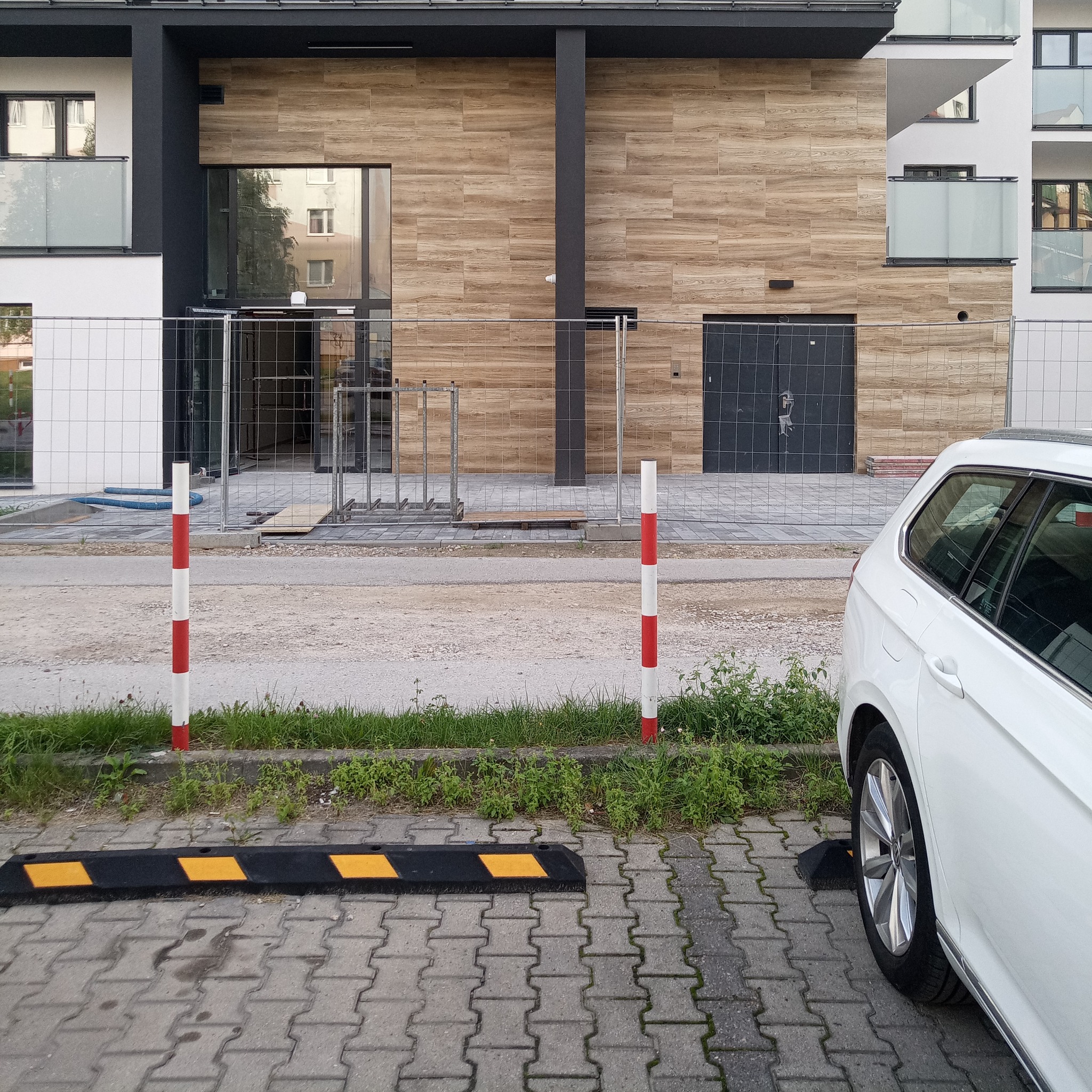 Nowoczesny budynek w trakcie budowy, zabezpieczony ogrodzeniem tymczasowym, z widocznymi elementami wykończeniowymi elewacji imitującej drewno i fragmentem białego samochodu osobowego.