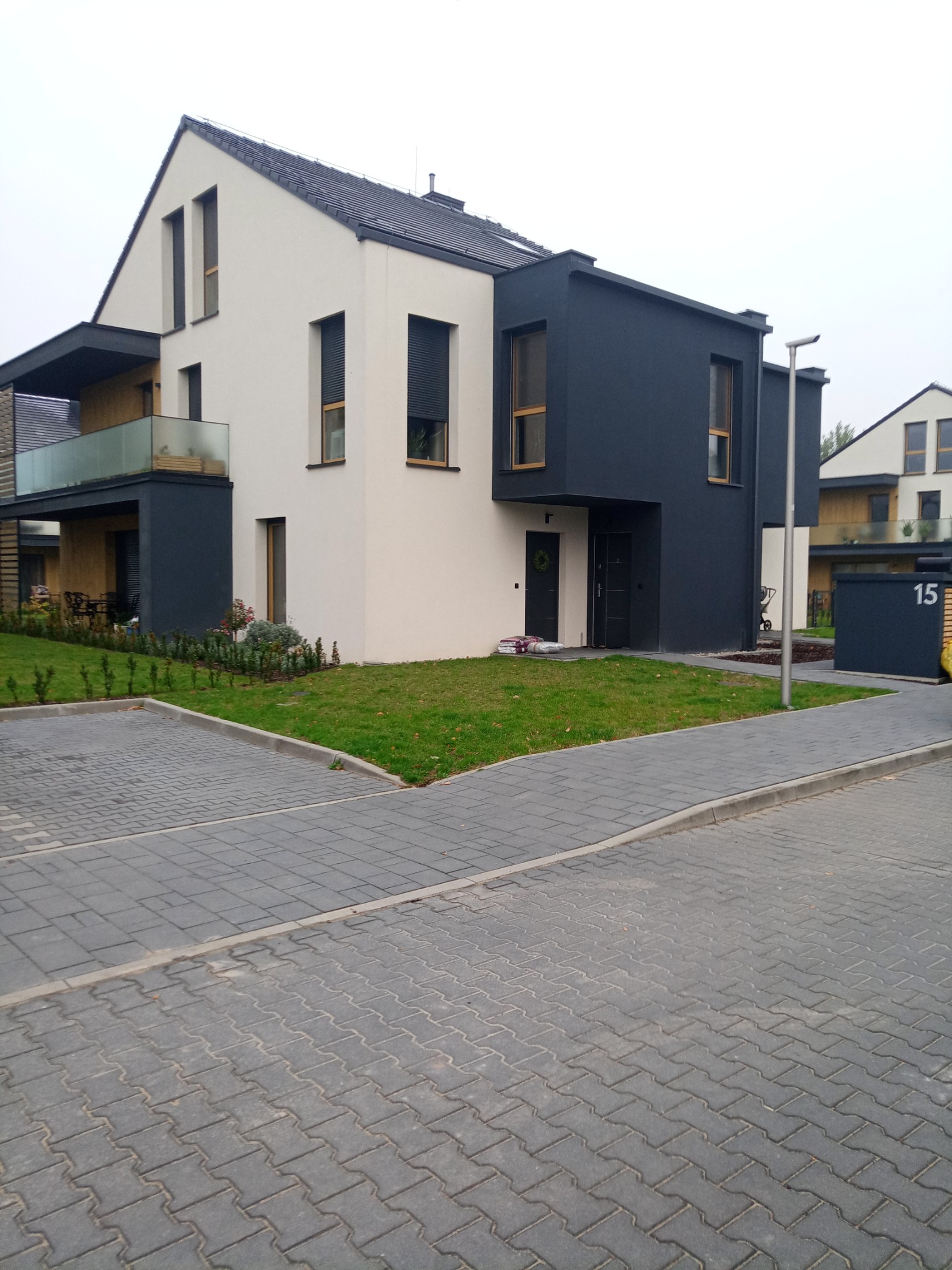 Nowoczesny dom dwukondygnacyjny z płaskim dachem, elewacja w kolorze bieli i antracytu, z oknami z drewnianą ramą, otoczony trawnikiem i brukowanym podjazdem.